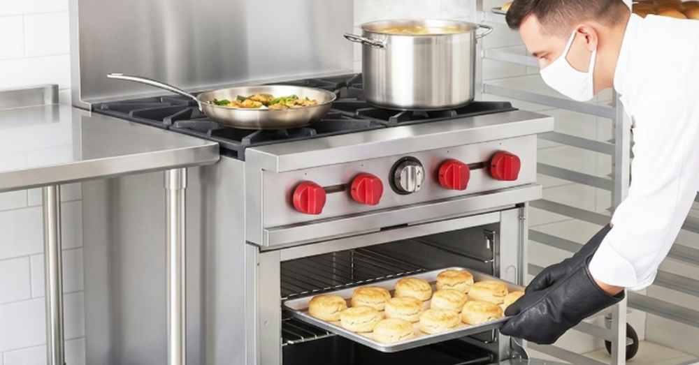 A chef wearing a white face mask removes a large tray of fresh biscuits from a small commercial oven.