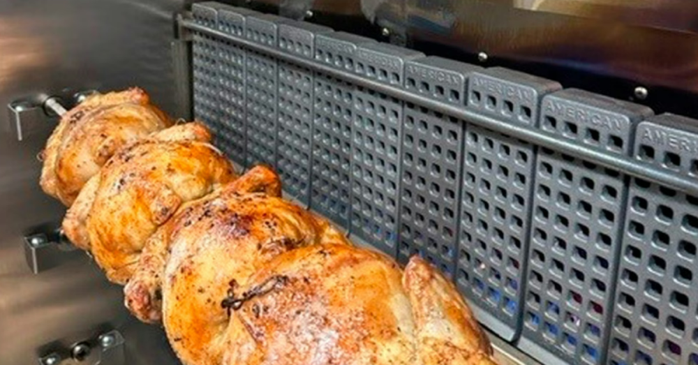 Four whole rotisserie chickens on a spit inside a rotisserie cooker. Another chicken is on a spit lower inside the oven.