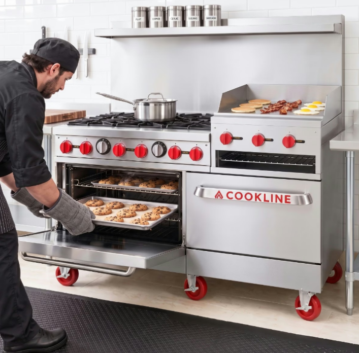 A chef removing cookies from an oven while pancakes, bacon, and sunny-side-up eggs cook on a nearby griddle.