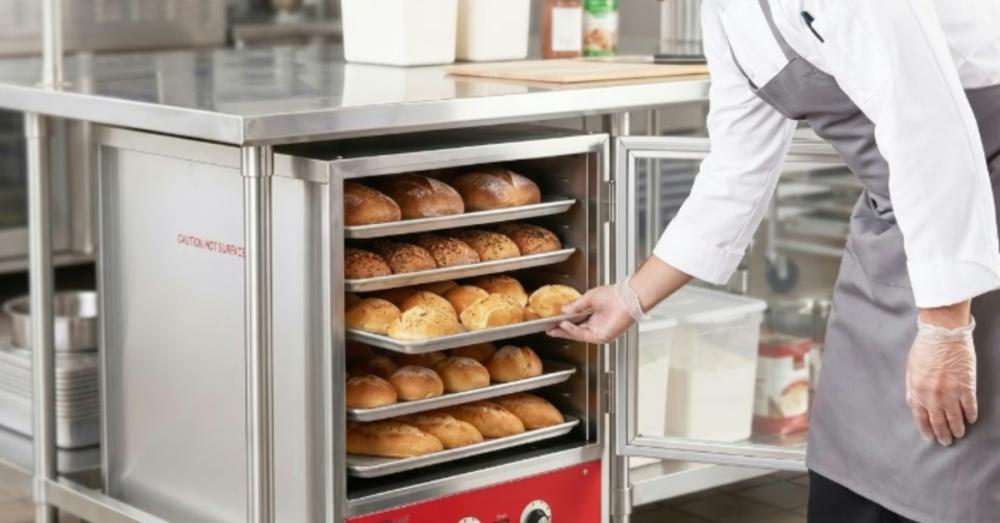 A baker bending over to remove a pan full of bread from a rack inside a small metal oven with a glass door.