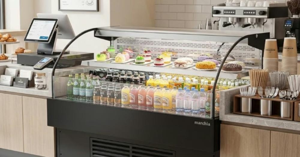 A short, wide commercial open air cooler full of products installed next to a cash register inside a shop.