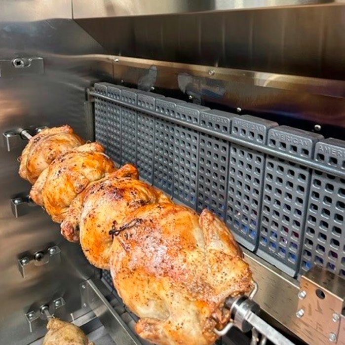 Four large chickens cooking inside a rotisserie oven. Another chicken is cooking on a spit underneath.