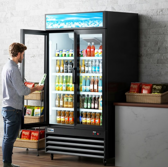 Customer removing a small glass bottle from a tall two-door merchandiser fridge in a convenience store.