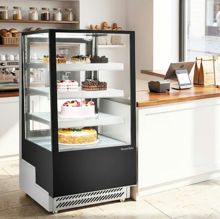 A large refrigerated bakery display case full of cakes. The display case sits beside the cash register.