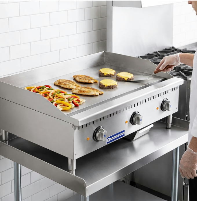 A chef cooking cheeseburgers, chicken breasts, and sliced peppers on a griddle in a commercial kitchen.